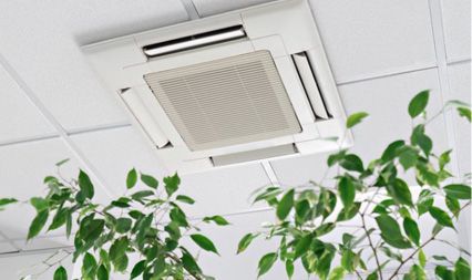A white ceiling-mounted HVAC cassette unit with vents, viewed from below with green plants in the foreground.