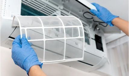 A person wearing blue gloves removes a white plastic mesh filter from a wall-mounted air conditioner.