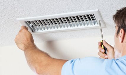 A person in a light blue shirt uses a screwdriver to install or remove a white rectangular ceiling air vent cover.