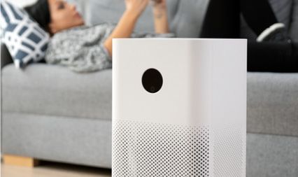 A white air purifier stands in the foreground, while a person reclines on a sofa in the blurred background.