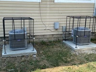 Two outdoor air conditioning units sit on concrete pads, each enclosed in a black metal security cage against a house wall.