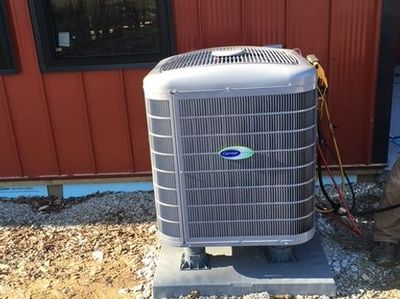 A grey Carrier air conditioning unit sits on a concrete pad outside a building with red metal siding.