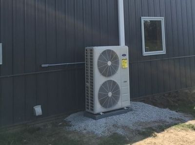 An outdoor HVAC unit on a gravel pad against a dark gray metal-sided building with a small window.