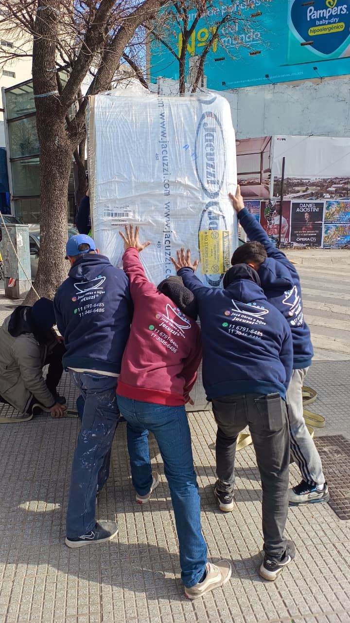 Cinco personas levantan un objeto rectangular grande y envuelto al aire libre en un día soleado.