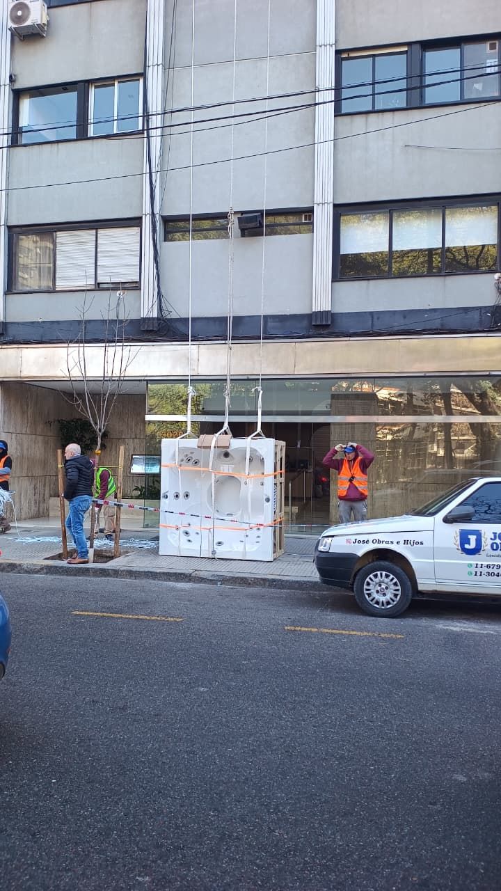 Exterior del edificio con una gran caja que bloquea la entrada, gente en la acera y un coche blanco en la calle.