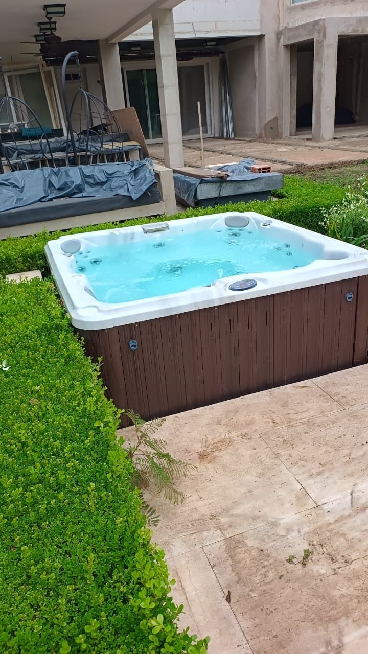 Jacuzzi con agua burbujeante junto a arbustos verdes en un patio trasero con construcción al fondo.
