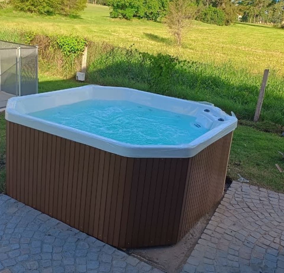Jacuzzi en un patio, revestimiento marrón, agua burbujeante. Césped verde y árboles al fondo.