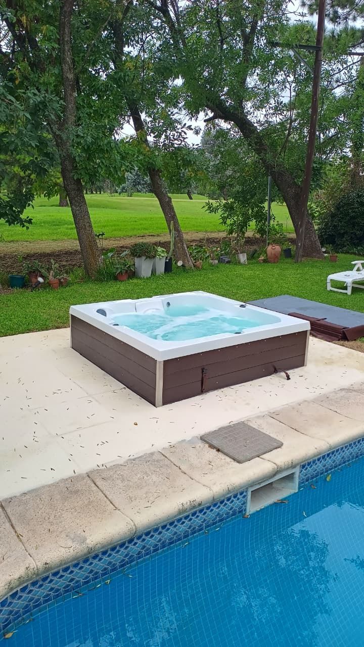 Jacuzzi junto a una piscina en un patio de hormigón, césped verde y árboles al fondo.