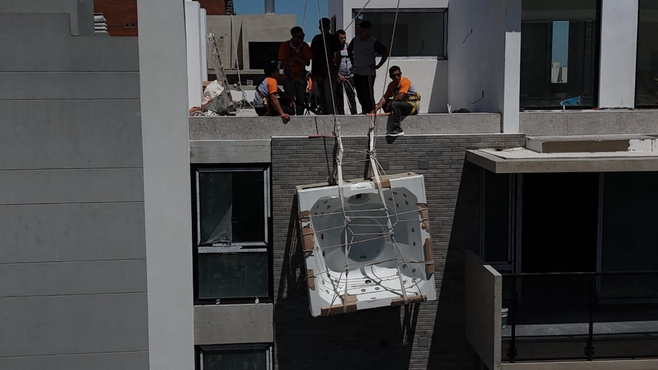 Trabajadores bajando una gran unidad de metal desde el tejado de un edificio alto, en un día soleado.