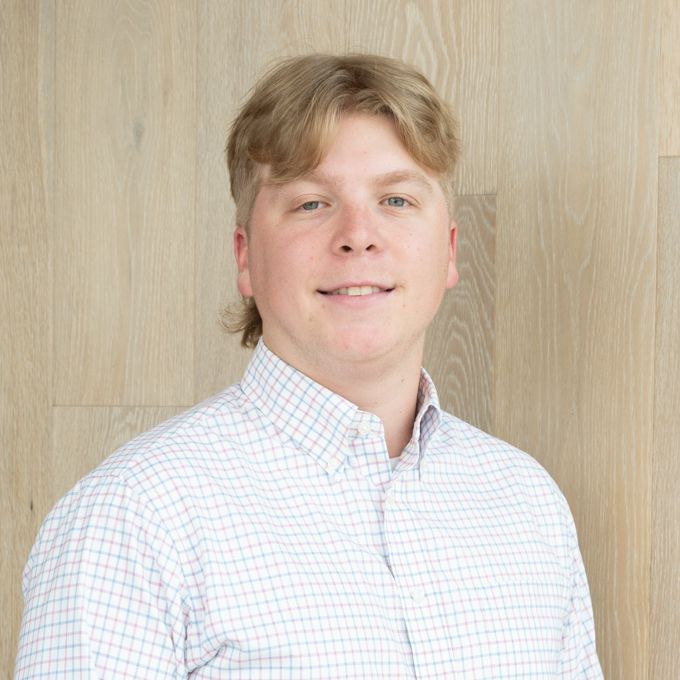 Person in a white checkered shirt standing against a light wood wall, looking at the camera.