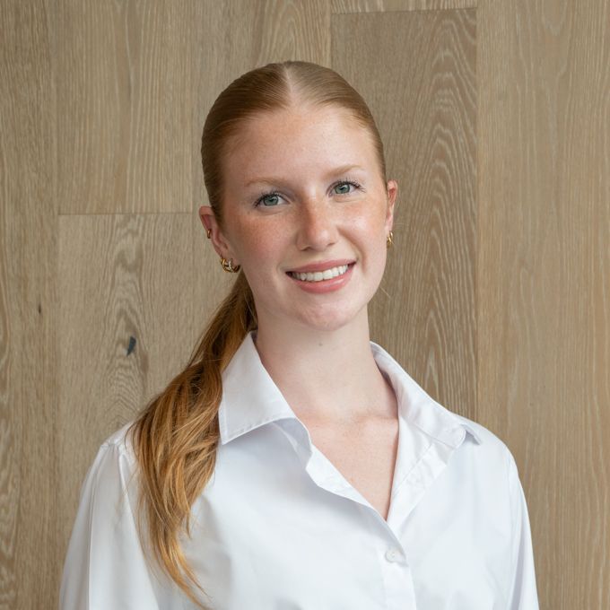 Smiling person in a white shirt standing against a light wood wall
