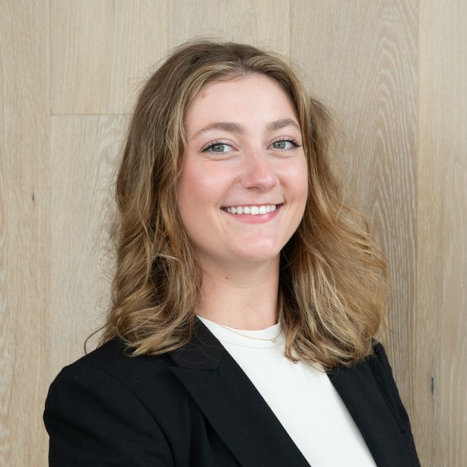 Smiling person in a black blazer and white top standing before a light wood wall