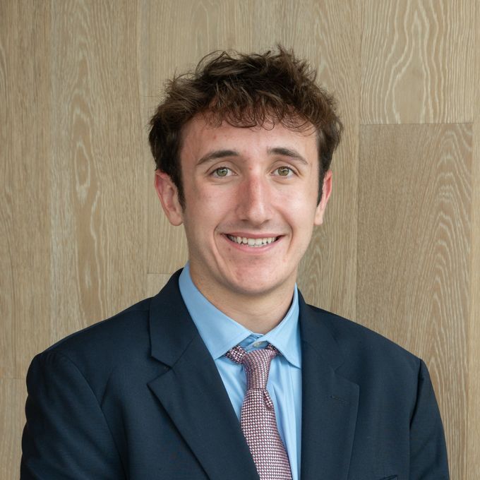 Smiling person in a navy suit and blue shirt against a light wood wall