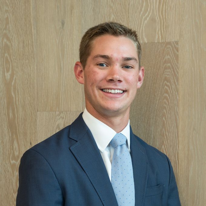 Smiling person in a navy suit and light blue tie against a wood-paneled wall