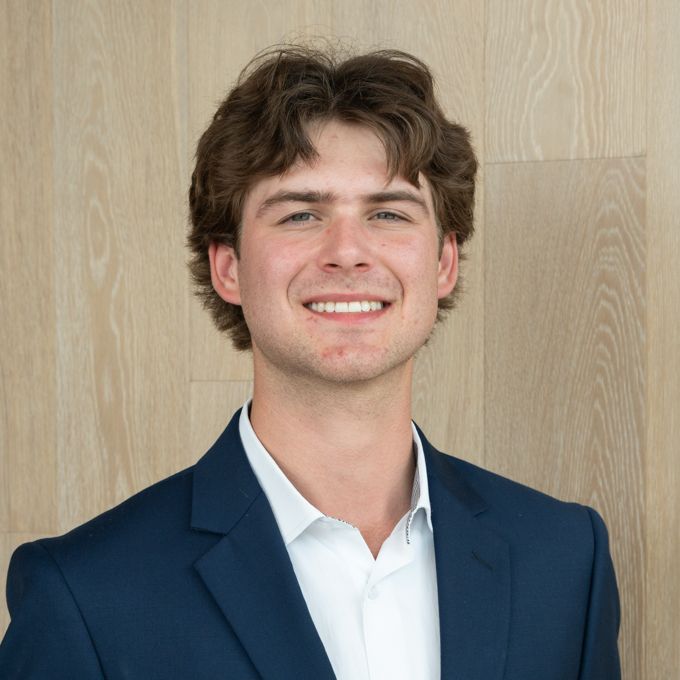 Smiling person in a navy blazer and white shirt against a light wood wall