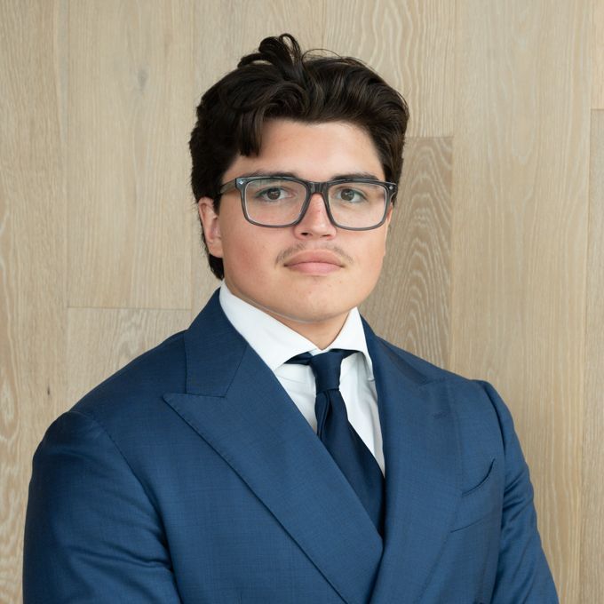 Portrait of a man in a blue suit, white shirt, and tie, standing against a light wood wall.
