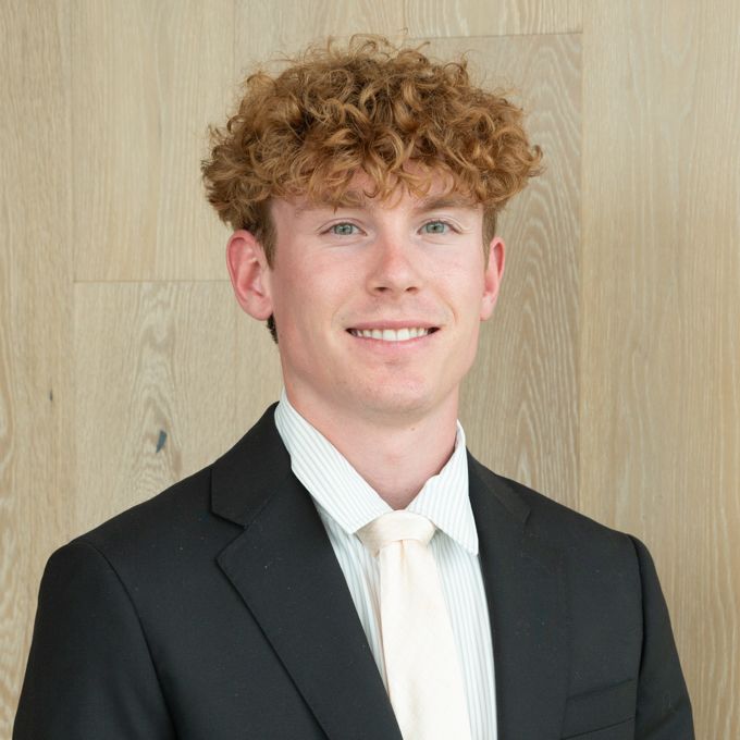 Portrait of a smiling person in a black suit and white tie against a light wood wall