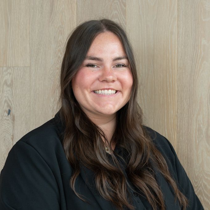 Smiling person with long dark hair in a black top against a light wood wall