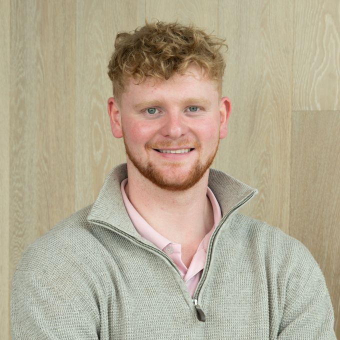 Smiling person in a light gray sweater standing against a wooden wall