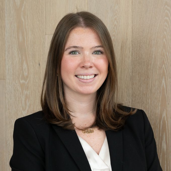 Smiling woman in black blazer and white top against a light wood wall