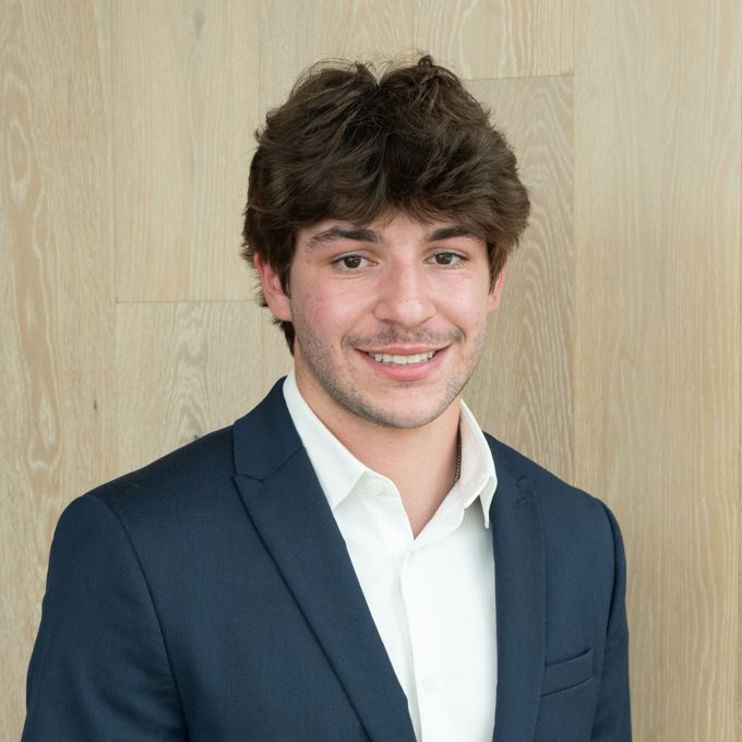 Smiling man in a navy blazer and white shirt standing against a light wood wall