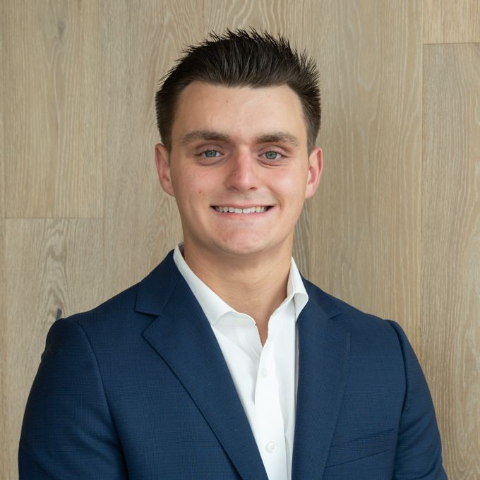 Smiling man in a navy suit and white shirt against a light wood wall