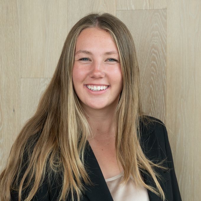 Smiling woman with long blonde hair in a black blazer against a light wood wall