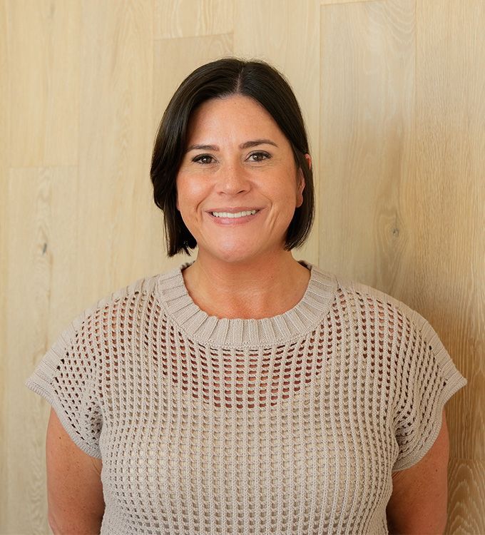 Smiling woman in a beige knit top standing against a light wood wall
