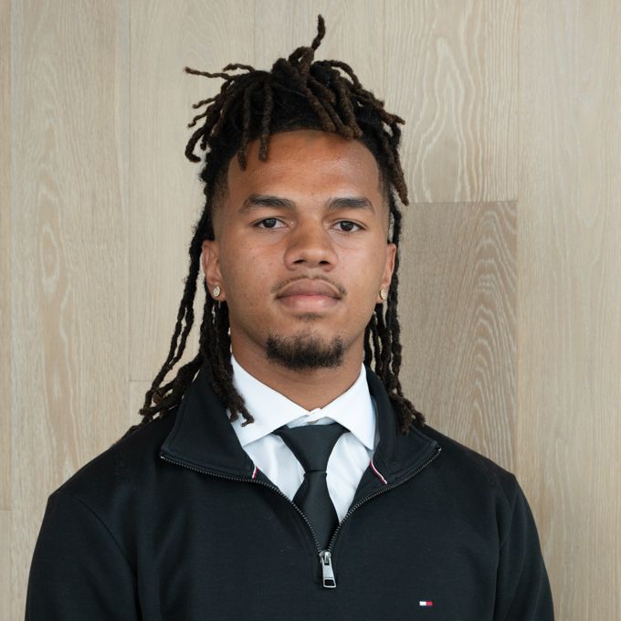 Portrait of a man in a black zip-up jacket, white shirt, and tie against a wood-paneled background