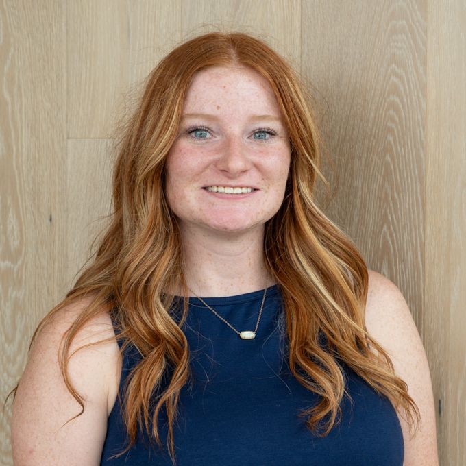 Smiling person with long red hair wearing a blue top against a light wood wall