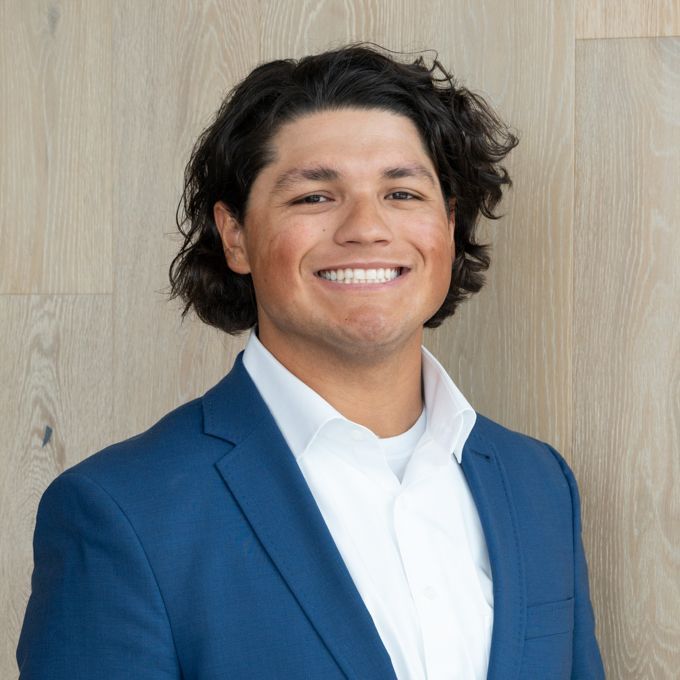 Smiling person in a blue suit jacket and white shirt against a light wood wall