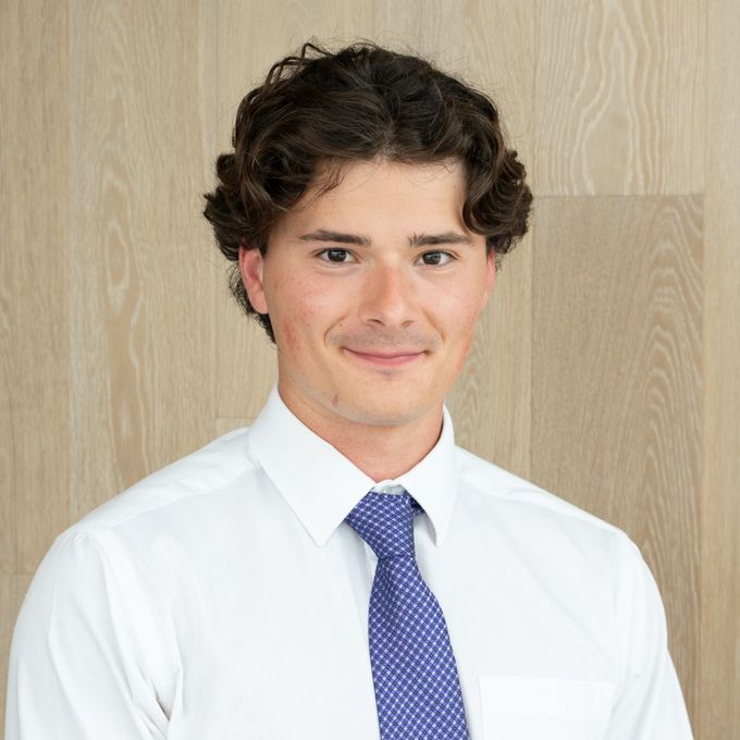 Smiling person in a white shirt and blue tie against a light wood wall