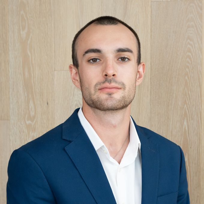 Man in a blue blazer and white shirt posing against a light wood wall