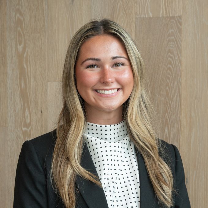 Smiling person in a black blazer and white polka-dot blouse standing before a wood-paneled wall