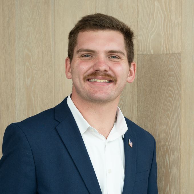 Smiling man in a navy blazer and white shirt standing against a wood-paneled wall