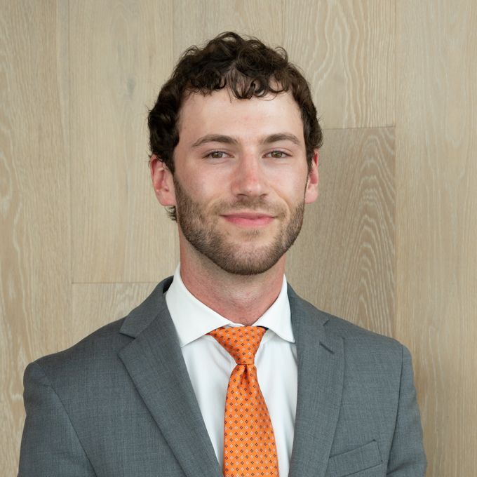 Portrait of a man in a gray suit and orange tie standing against a light wood wall