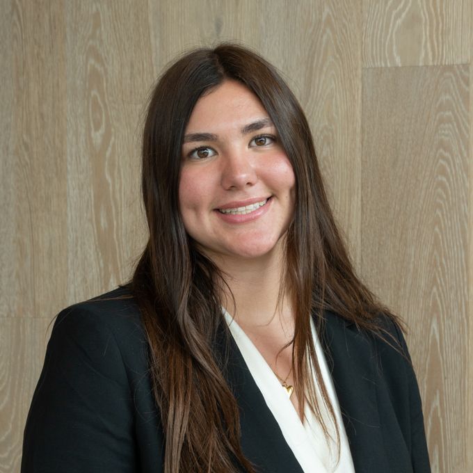 Smiling person in a black blazer and white blouse against a wood-paneled wall