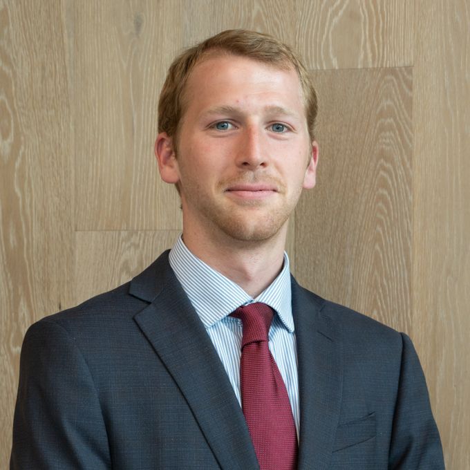 Person in a dark suit and red tie posing against a wood-paneled wall.