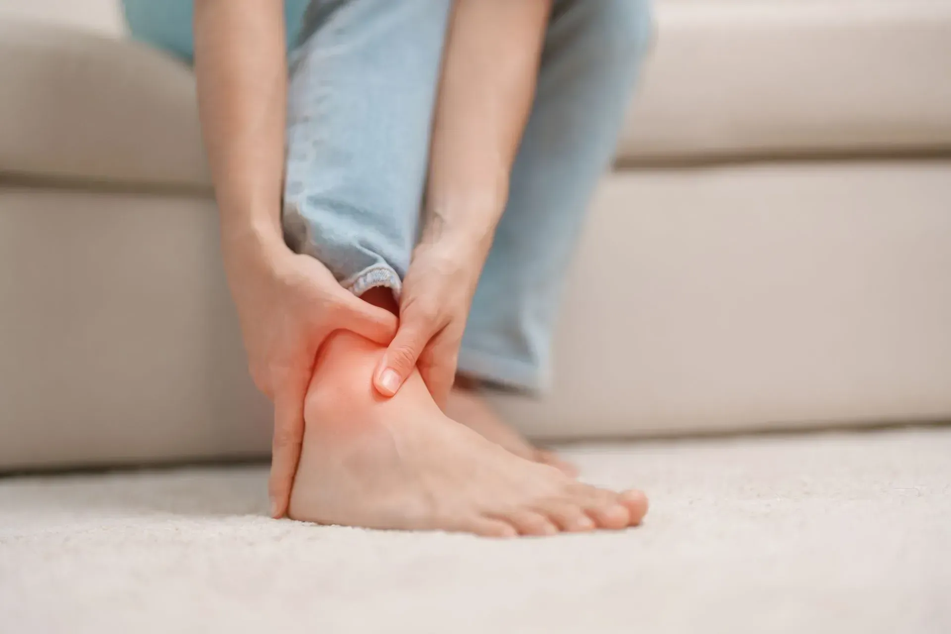 A person holding their ankle with a red, highlighted area indicating pain, while sitting on a light-colored surface.