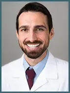 Headshot of a person with a beard, wearing a white lab coat, light blue collared shirt, and a dark tie.