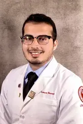 A person in a white lab coat and glasses, smiling, set against a grey background.