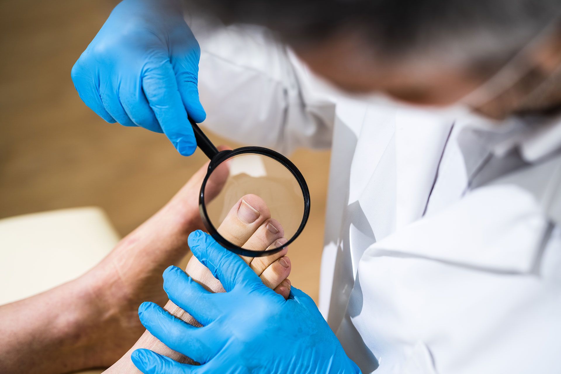 A professional wearing a white coat and blue medical gloves uses a magnifying glass to examine a patient’s toenails.