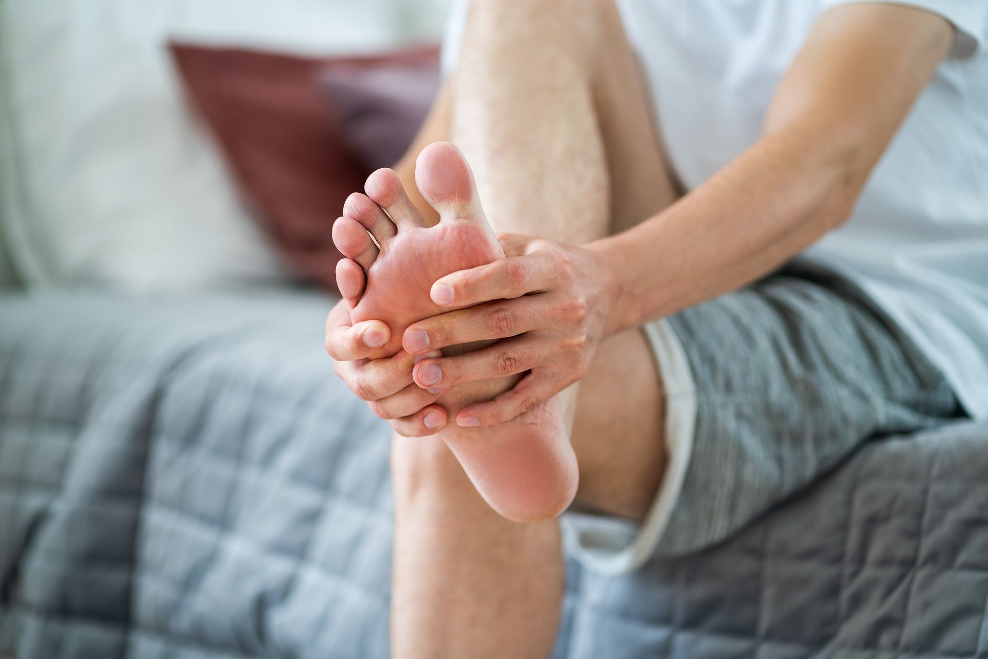 A person sits on a bed, holding and massaging their bare foot with both hands.