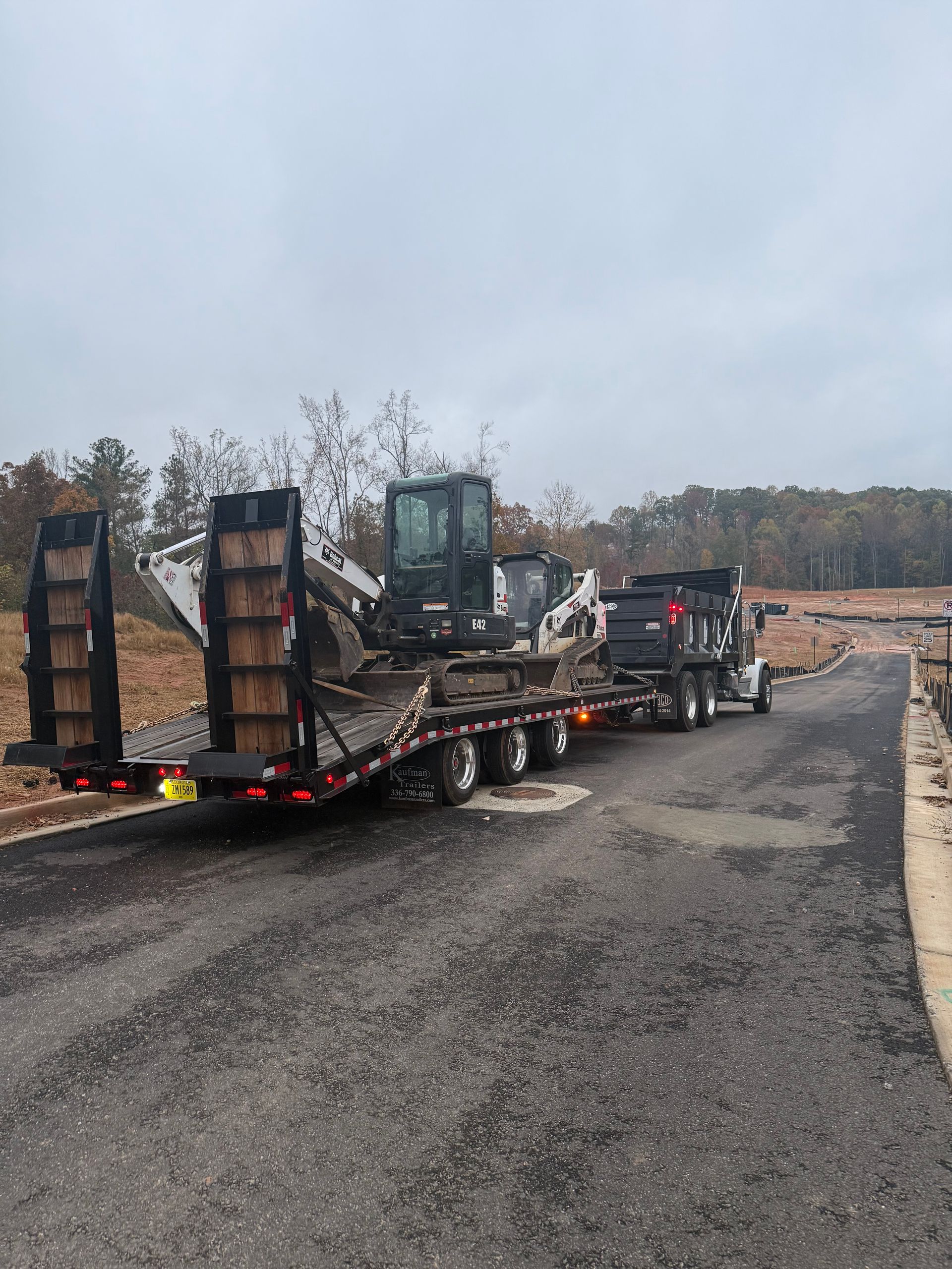 A flatbed trailer carrying a white excavator and a black skid steer, attached to a truck on a paved road near a wooded area.
