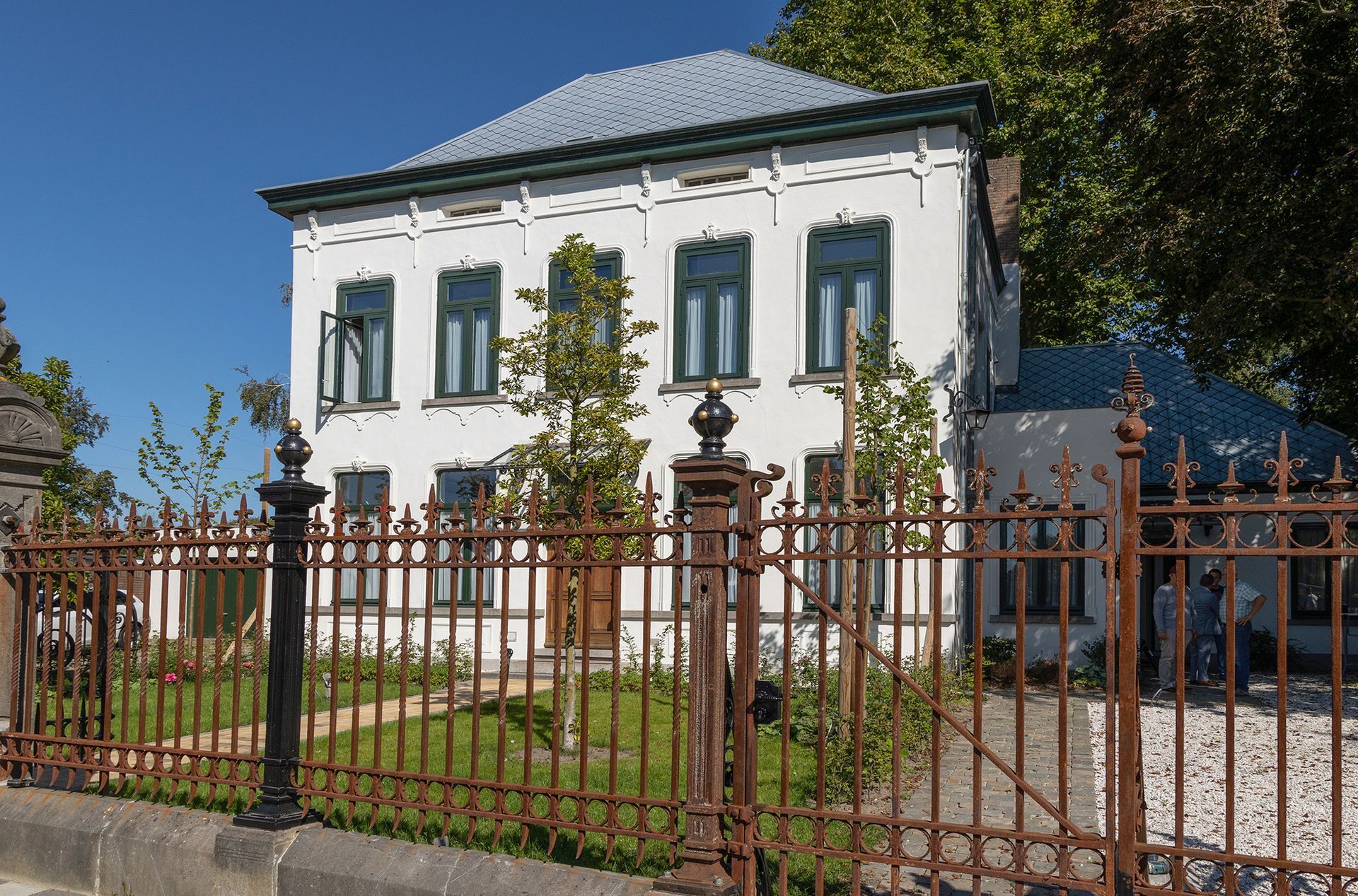 Een wit gebouw met groen omlijste ramen, een blauw dak en een bruin metalen hek op een zonnige dag.