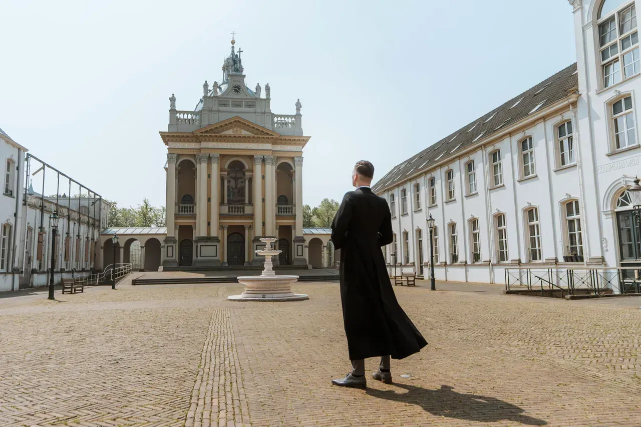 Een persoon in een zwart gewaad staat en kijkt naar een wit gebouw met een gedetailleerde gevel op een binnenplaats.