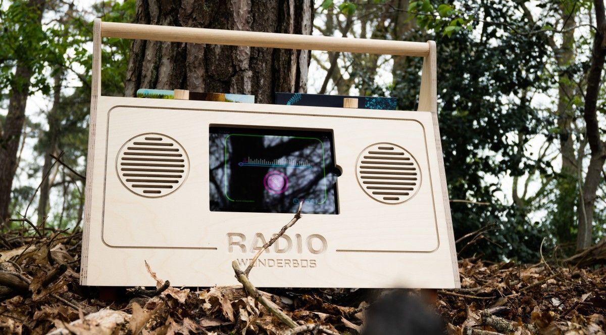 Een houten, boombox-achtig apparaat met een scherm staat in een bos, voor een boom.