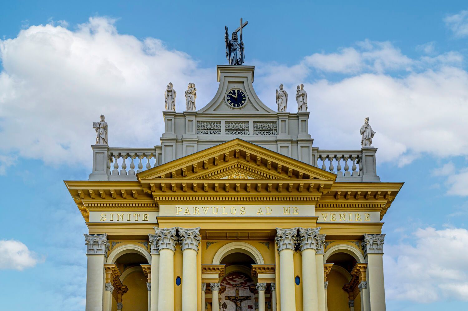 Een sierlijk geel-wit gebouw met zuilen, sculpturen en een klok tegen een blauwe hemel.