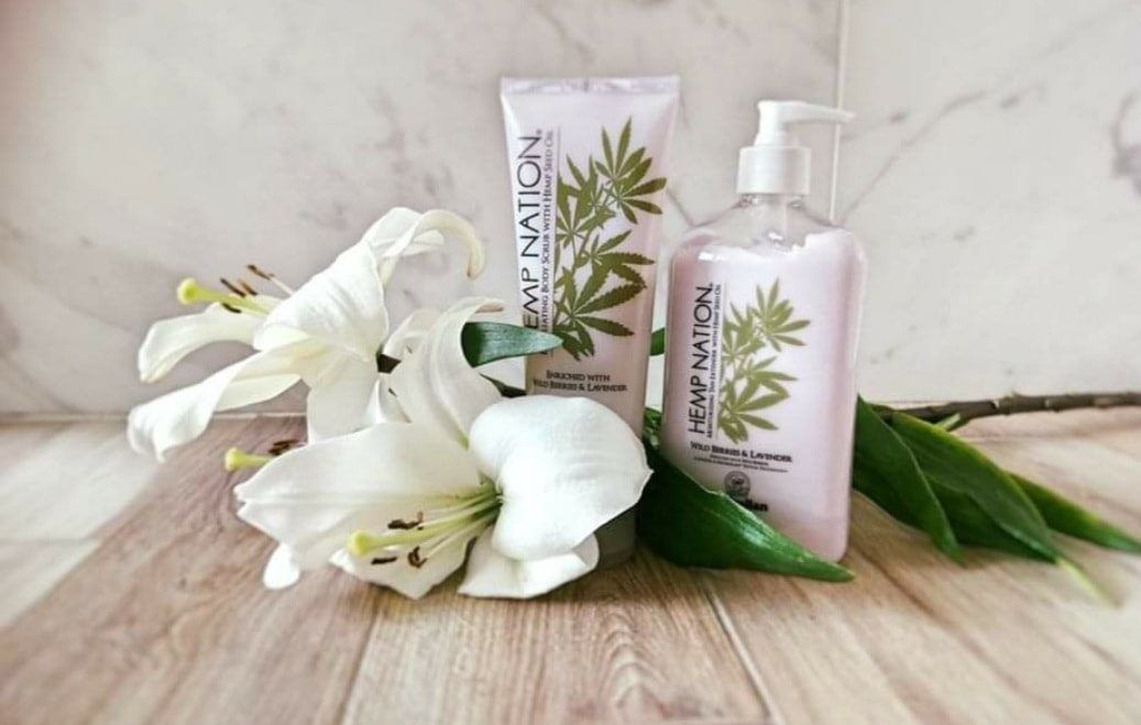 Due prodotti per la cura della pelle Hemp Nation con gigli bianchi su superficie in legno.