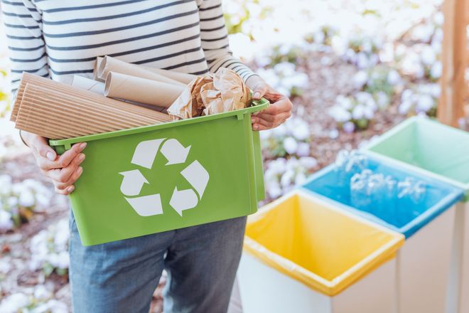 Person holding a green recycling bin filled with paper, with other bins visible in a bright outdoor setting.